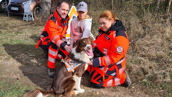 Ein Vater streichelt mit seinen Kindern den Familienhund. Auf dem Geschirr des Hundes steht: Rettunghund.