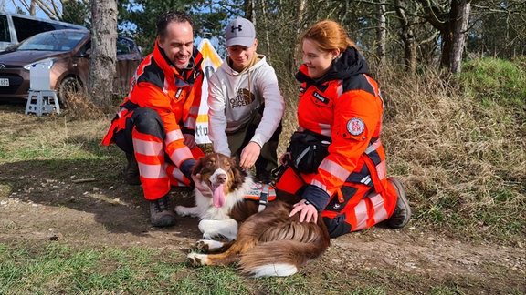 Ein Vater streichelt mit seinen Kindern den Familienhund. Auf dem Geschirr des Hundes steht: Rettunghund.