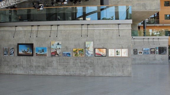 In einem lichtdurchfluteten Foyer hängen an Betonwänden - wie in einer Galerie - verschiedene Gemälde iund Fotografien dicht beieinander