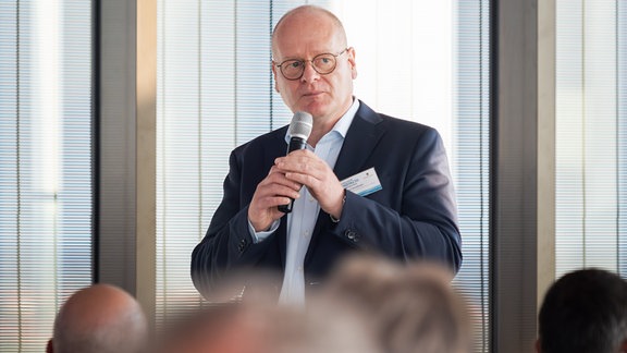 Ein glatzköpfiger Mann steht mit einem Mikrofon in der Hand vor Zuhörern.