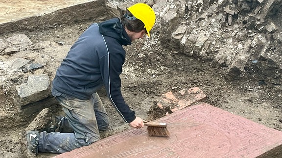 Ein Mann mit Helm kniet neben einer Grabplatte und säubert sie.