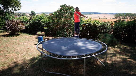 Ein kleines Mädchen hüpft auf einem Trampolin in einem umzäunten Garten vor einem weiten Feld