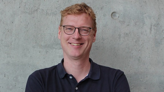 Ein großer Mann mit Locken und Brille steht vor einer Betonwand.