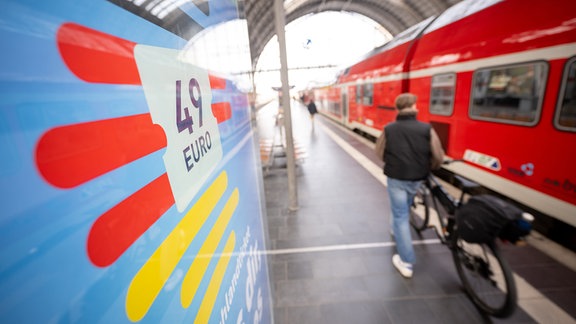 Ein Mann geht am Hauptbahnhof mit seinem Fahrrad zwischen der Werbung für das Deutschlandticket und einer Regionalbahn vorbei.