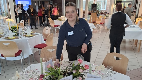 Eine junge Frau steht an einem runden gedeckten Tisch und lächelt in die Kamera.