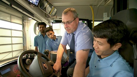 Ein Mann zeigt drei jungen Männern etwas am Steuerrad eines Busses.