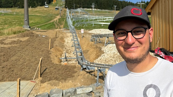 Ein junger Man mit Basecap steht vor einem Berghang mit einer Sommerrodelbahn.