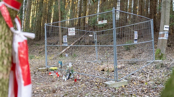 Eine mit Bauzäunen abgesperrte Stelle im Wald, an der Blumen, Kerzen und Bilder niedergelegt sind.