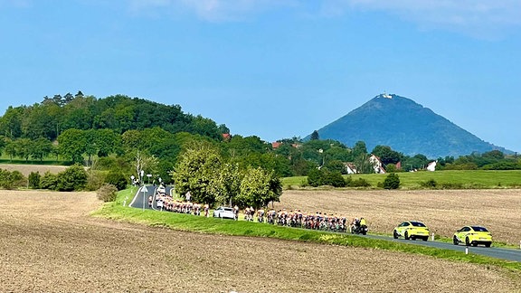 Auf einer Landstraße fahren Radsportler und Begleitfahrzeuge eines Etappenrennens. Dahinter ein steiler Berg mit Burg.