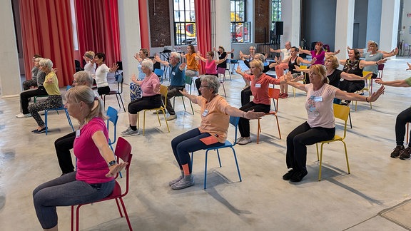 Blick in einen großen Raum, in dem mehrere ältere Menschen auf bunten Stühlen sitzen und eine gemeinsame Choreografie aufführen