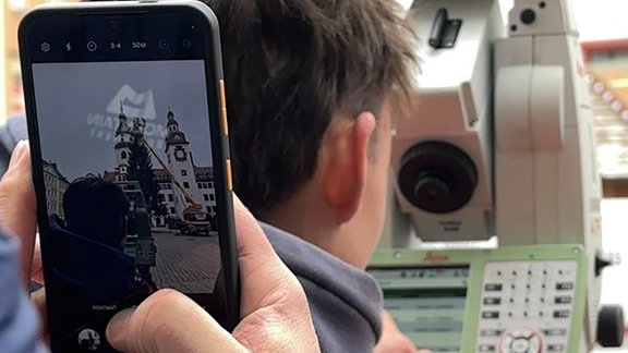 Es ist von hinten ein Mann mit roter Jacke zu sehen, der ein Vermessungsgerät bedient. Dahinter steht ein Mann mit blauer Jacke, der ein Foto vom Mann mit roter Jacke macht. Das ist im Handydisplay zu sehen und im Hintergrund ist dort auch zu erkennen, was vermessen wird: der Weihnachtsbaum auf dem Marktplatz in Chemnitz.  