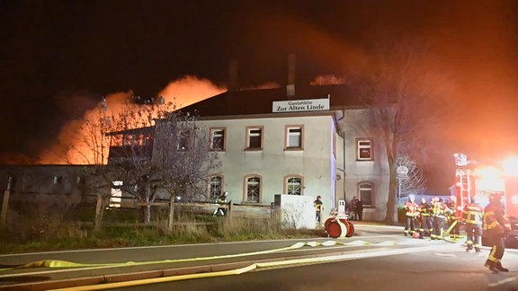 Ein Haus, Feuerschein im Hintergrund, Feuerwehrleute auf der Straße