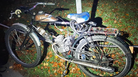  Ein Fahrrad, an dem nachträglich ein Mopedmotor angebaut wurde, lehnt an einem Zaun. 