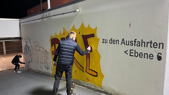 (Zwei Männen sprühen an einer weißen Wand ein Graffiti.)