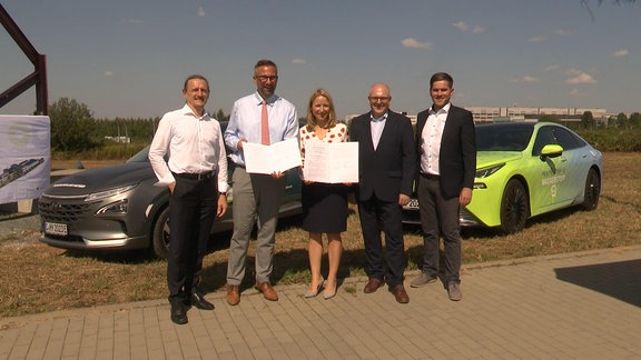 Fünf Personen posieren in einem "Familienfoto" vor zwei Autos auf dem Baugelände des Wasserstoffzentrums in Chemnitz, darunter Wirtschaftsminister Martin Dulig, SPD.