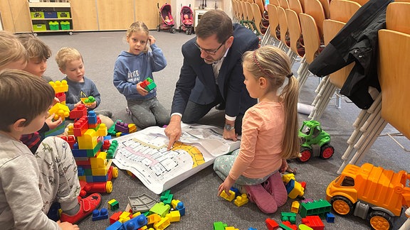 Der Bürgermeister von Steinberg, Andreas Gruner, und Kinder der Rothenkirchener Kita "Steinbergwichtel" hocken im Kreis um den Bauplan für die neue Kita.