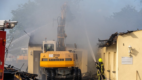 Ein Bagger räumt Trümmer weg, während ein Feuerwehrmann löscht.