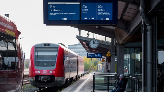 Eine Anzeigetafel am Bahnsteig mit den Fahrzielen Lichtenfels, Hof und Bamberg und zwei haltenden Zügen der Deutschen Bahn