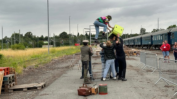 Ein hochkant gestelltes Absperrgitter wird benutzt, um einen hohen Zaun anzudeuten. Über diesen steigt gerade eine Frau, der ein Kinderwagen gereicht wird. 