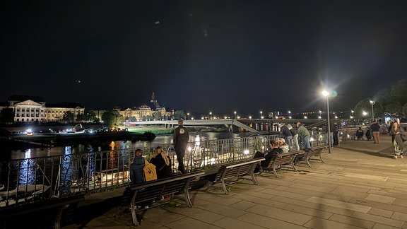 An einer Brücke im Dunkeln sind Bagger zu sehen. Vom gegenüberliegenden Ufer aus betrachten einige Menschen den Schauplatz am späten Abend.