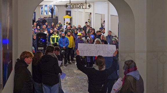 Eine größere Gruppe von Menschen steht in einem Foyer. Sie tragen Jacken und Fahrradhelme und schauen zu einer Person die mit einem Banner vor ihnen steht.