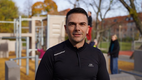 Ein Mann im dunklen Trainingsanzug schaut freundlich in die Kamera. Im Hintergrund eine Art Klettergerüst.