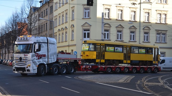 Ein weißer Sattelzug biegt mit einer gelben Straßenbahn beladen um eine Straßenecke. 