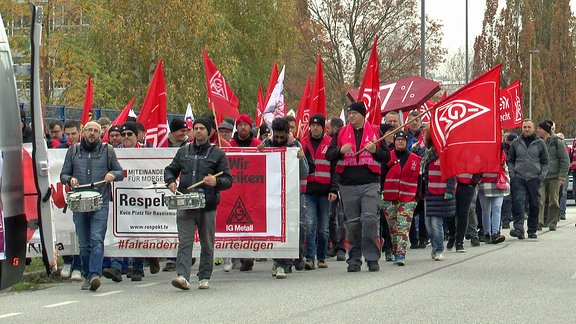 Beschäftgte vonMetallbetrieben gehen bei einer demo hinter Gewerkschaftern her, die Fahnen und Trommeln tragen.