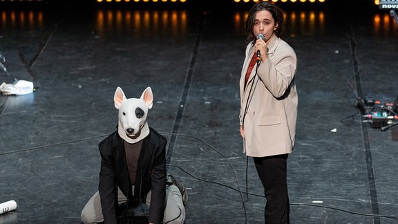 Zwei Menschen auf einer Bühne, eine Schauspielerin steht mit einem Mikrofon in der Hand, ein Schauspieler kniet und hat eine Hundemaske auf dem Kopf.