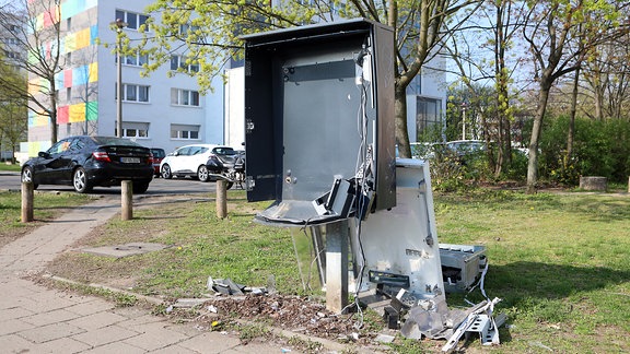 Am Rande eines Parkplatzes vor einem Plattenhochhaus steht ein Zigarettenautomat, der nur noch aus einer leeren Verschalung besteht. Die untere Seite ist ausgebeult, Kabel hängen lose herum. Hinter dem Automat liegt das ehemalige Innenleben auf der Wiese