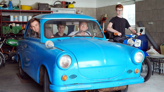 Drei Jungen sitzen in einem blauen Auto. Ein anderer steht mit seinem blauen Moped daneben.