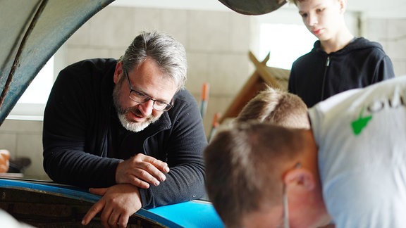 Ein älter Mann mit Vollbart und Brille sowie drei Jungs schauen den Motor eines Autos an