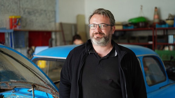 Ein älterer Mann mit Vollbart und Brille steht vor einem blauen Auto