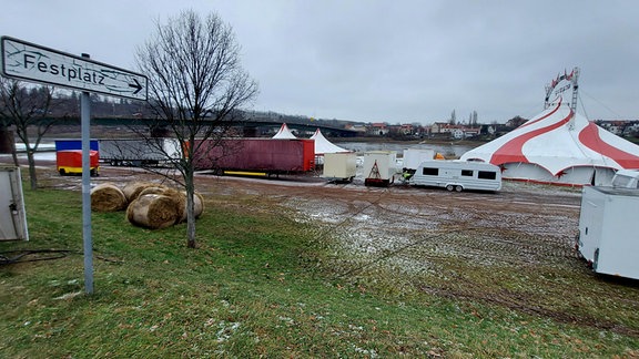 Auf einer Wiese steht ein Schild, das den Schriftzug "Festplatz" trägt. Auf dem Platz dahintersteht stehen ein Zirkuszelt und Wohnwagen.