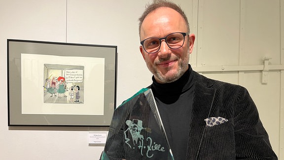 Ein Mann mit schwarzem Jackett und Brille hält eine gläserne Skulptur in der Hand. 