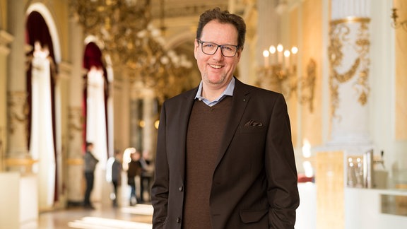 Peter Theiler, ein Mann in braunem Anzug und braunem Pullover darunter, Brille und braunem Haar steht lachend mit den Händen in den Hosentaschen in einem historischen Foyer.