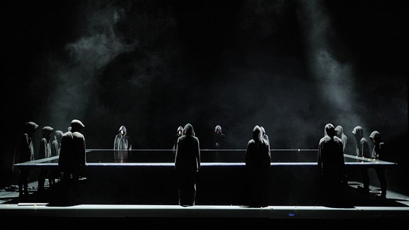 Szene aus dem Tanztheaterstück "Wonderful World": Menschen in schwarzer Kleidung mit Kapuze stehen auf einer Bühne um einen großen Tisch.