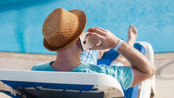 Mann liegt im Liegestuhl am Pool und telefoniert.