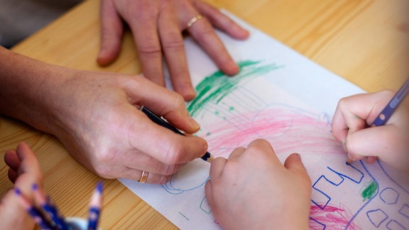 Die Hände einer Kindergaertnerin und eines Mädchens sind beim gemeinsamen Malen in einer Kita zu sehen.