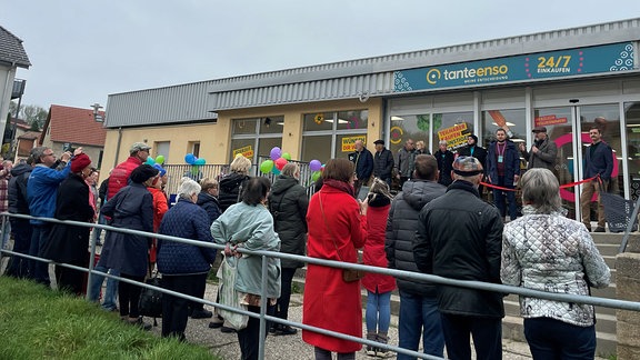  In Kohren-Sahlis stehen Menschen vor einem Supermarkt und verfolgen, wie das Rote Band zur Eröffnung durchgeschnitten wird.