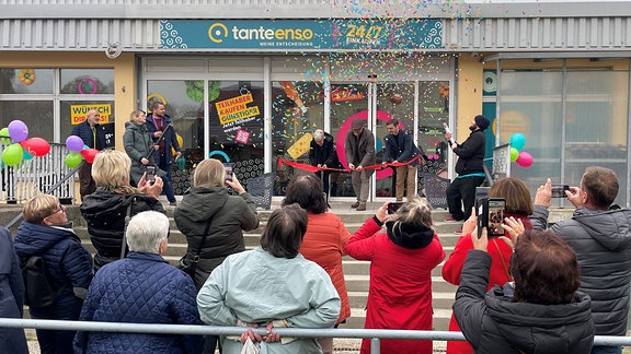  In Kohren-Sahlis stehen Menschen vor einem Supermarkt und verfolgen, wie das Rote Band zur Eröffnung durchgeschnitten wird.