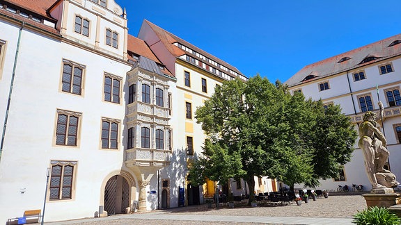 Ein weißes Renaissanceschloss steht in der Sonne. Im Hof steht ein grüner Baum.