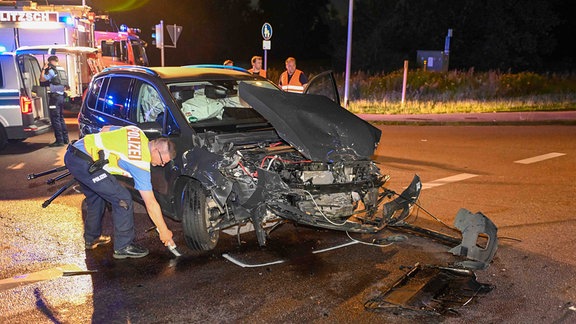 Ein Auto mit total demolierter Vorderfront steht auf einer Straße. Ein Verkehrspolizist markiert mit Kreide Stellen, die den Ablauf eines Verkehsunfalls darstellen.