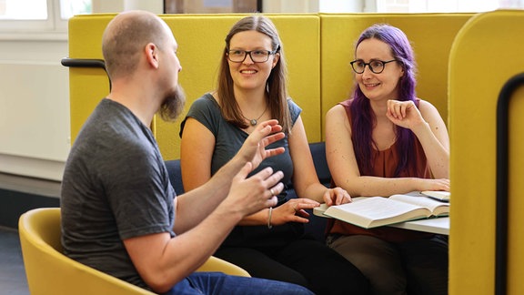 Die Vorstandsmitglieder des Vereins "Break the Fake": Drei Menschen sitzen an einem Tisch auf gelben Sitzmöbeln und besprechen sich.