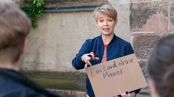 Eine Frau mit hellen, kurzen Haaren und blauem Blazer hält einen Stift und ein Pappschild mit der Aufschrift "Ein Land ohne Mauer" vor ihrem Körper. Sie steht vor einer alten Kirchenmauer.