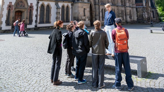 Eine Frau mit hellen, kurzen Haaren, blauem Blazer und Jeans steht auf einer steinernen Bank, ihr gegenüber stehen mehrere Personen. Hinter der Gruppe ist eine alte Kirche zu sehen.