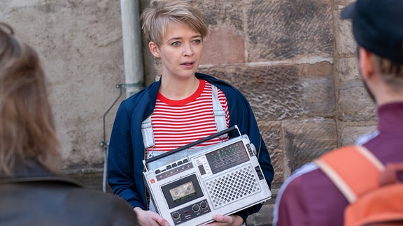 Einer Frau mit hellen, kurzen Haaren, blauem Blazer und Jeans hängt ein altes Radio um den Hals. Im Hintergrund ist ein altes Kirchengebäude zu sehen.