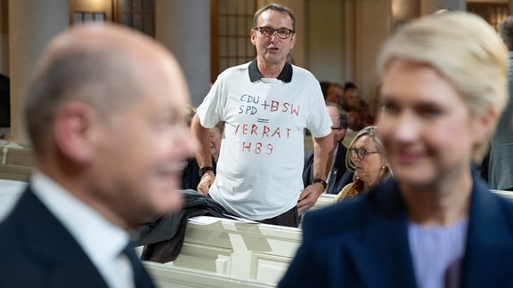 Ein Mann trägt vor Beginn einer Andacht in der Nikolaikirche hinter Bundeskanzler Olaf Scholz (SPD, l), und Manuela Schwesig (SPD), Bundesratspräsidentin und Ministerpräsidentin von Mecklenburg Vorpommern, ein T-Shirt mit der Aufschrift "CDU SPD + BSW = Verrat H 89". Anlass ist der Jahrestag der friedlichen Revolution vor 35 Jahren.