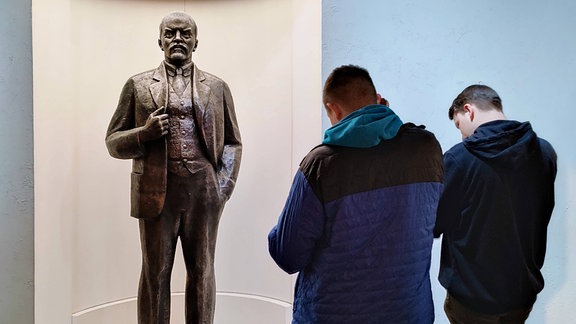 Jugendliche stehen vor einer Statue von Lenin, die in einer weißen, runden, futuristischen Vitrine im Museum Zeitgeschichtliches Forum leipzig steht