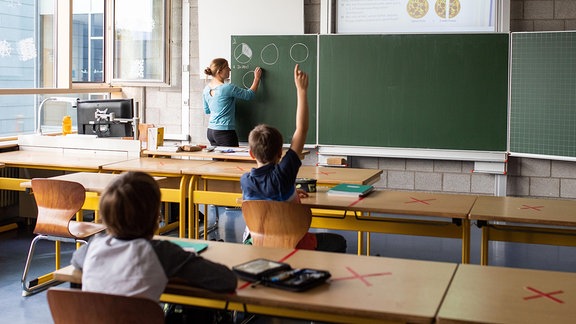 Schulkinder sitzen an ihren Tischen, während eine Lehrerin an der Tafel unterrichtet